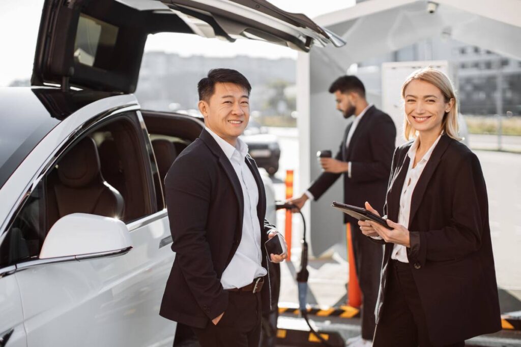 Relaxed smiling multiracial business colleagues standing at e-car charging point with tablet and mobile in hands. Indian man holding takeaway cup and getting auto filled up with electricity behind.