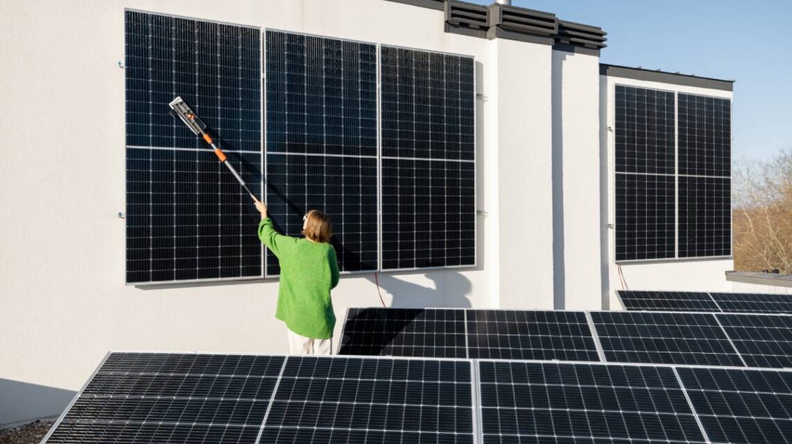 Woman washing solar panels with a brush on rooftop of her household on sunny day. Solar station care and maintenance concept