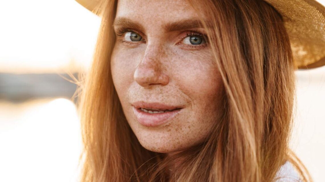 Image of pleased ginger woman in hat posing and looking at camera while walking on promenade