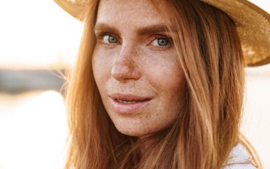 Image of pleased ginger woman in hat posing and looking at camera while walking on promenade